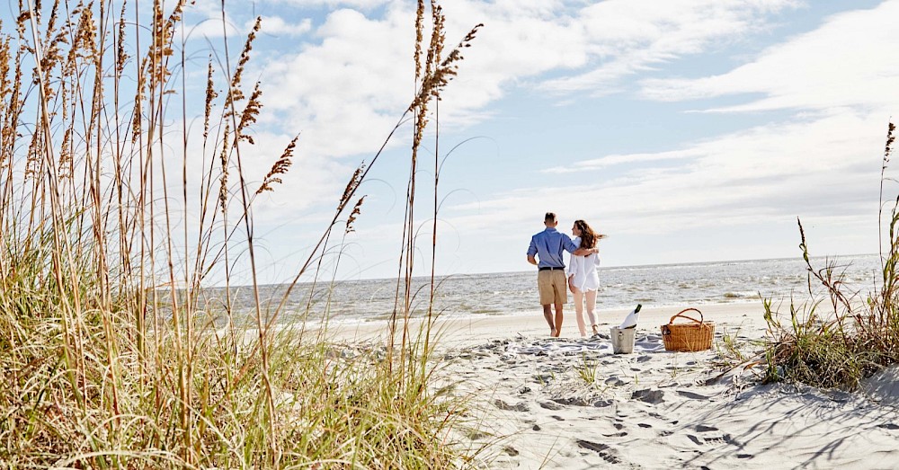 A couple walks hand in hand toward the sea on a sandy beach, with tall dune grasses in the foreground and a picnic basket nearby, enjoying a coastal stroll.