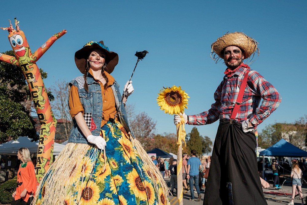 A sunny outdoor festival with costumed performers: a woman in a sunflower dress, a man in overalls and a hat, and a tall scarecrow figure in the background, posing cheerfully. End.