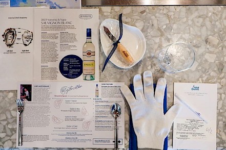 Wine tasting setup on marble table: menu, two spoons, a glass, glove, small plate with cheese, blue napkin, and a bottle of white wine.