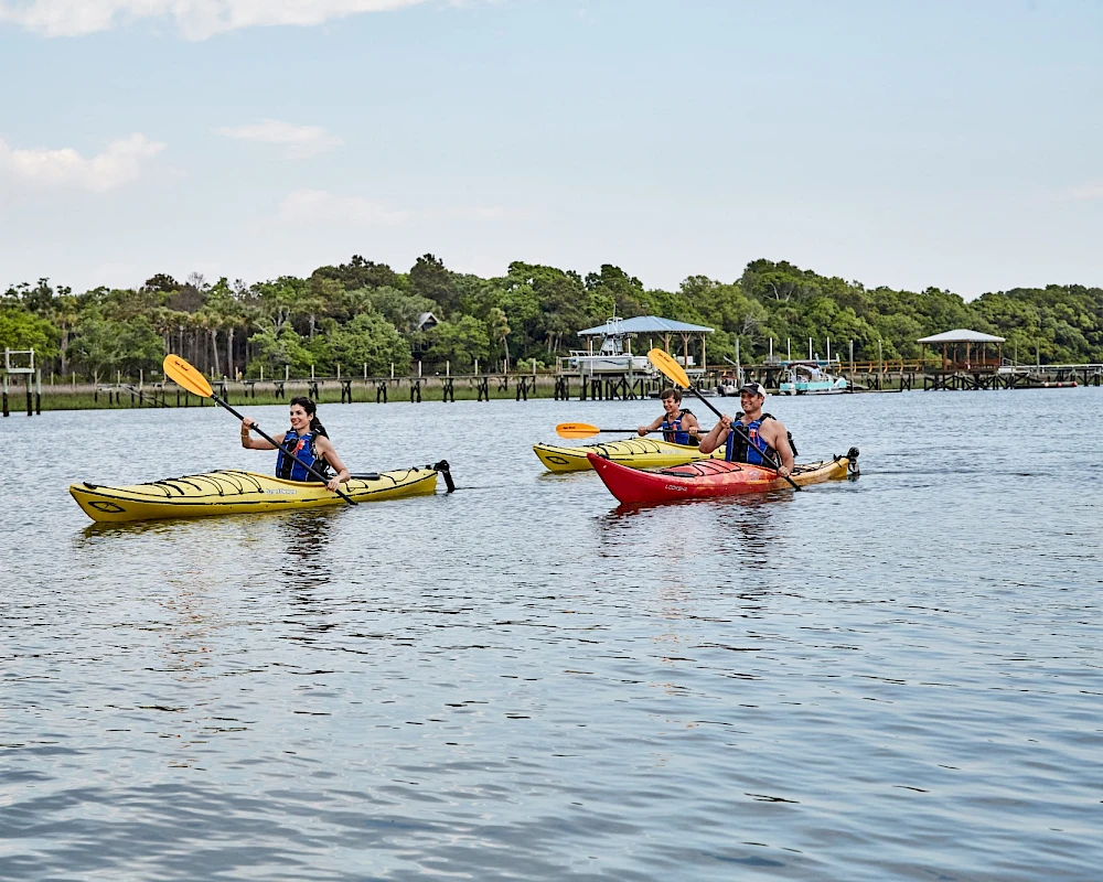 Two people kayak on calm water near a shoreline with docks and trees, paddling in yellow and red boats, sunny day.