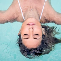 A person floating upside down in a turquoise pool, wearing a white swimsuit strap, with eyes closed and hair spread in the water.