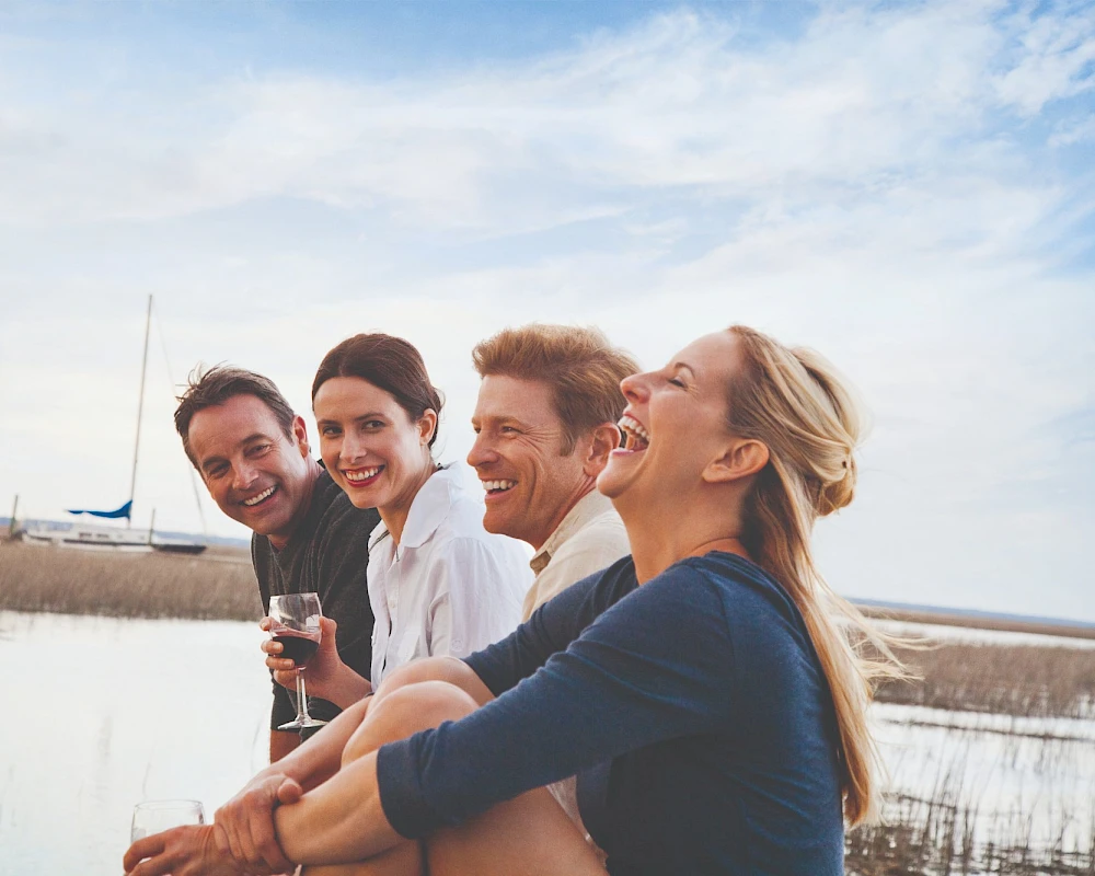 A group of four friends posing by a lakeside, smiling and enjoying drinks, with a boat in the background.