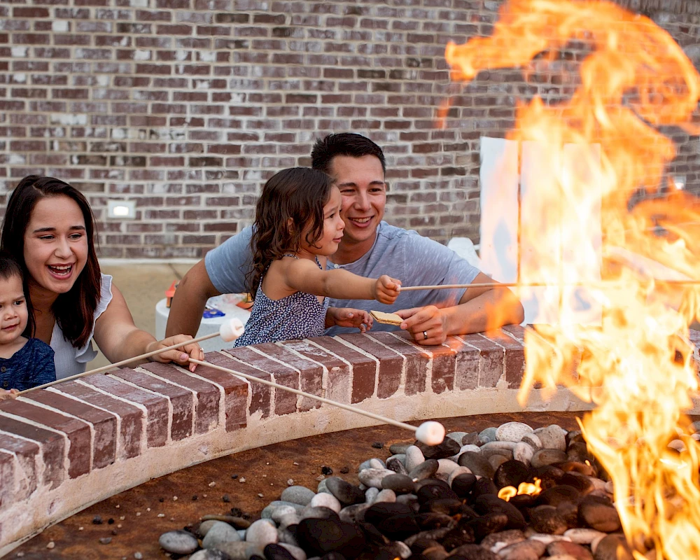 A family peers over a brick fire pit, smiling and reaching toward tall flames; kids hold hands while parents supervise the glow.