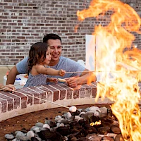 A family peers over a brick fire pit, smiling and reaching toward tall flames; kids hold hands while parents supervise the glow.