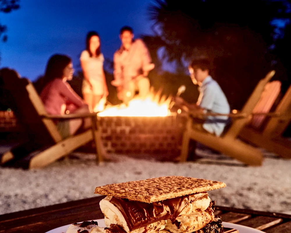 A cozy bonfire scene with friends around a fire pit at dusk, while a stack of grilled sandwiches sits on a plate in the foreground.