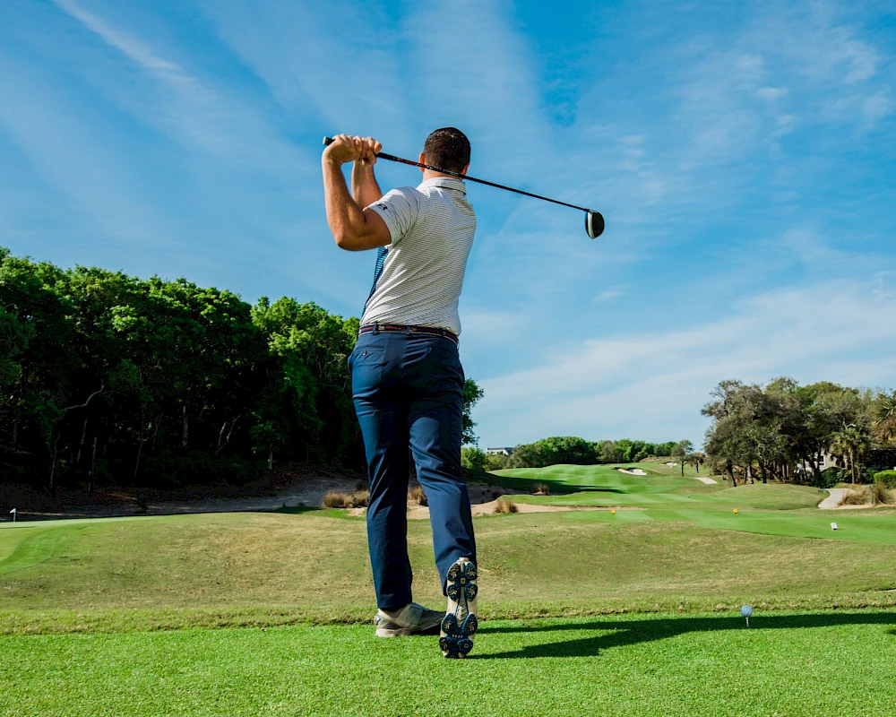 A golfer in mid-swing on a sunny golf course, blue sky, green fairway, and trees lining the course.