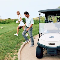 A couple playfully jog along a golf path beside a white golf cart on a sunny course, ready to hit their next shot.