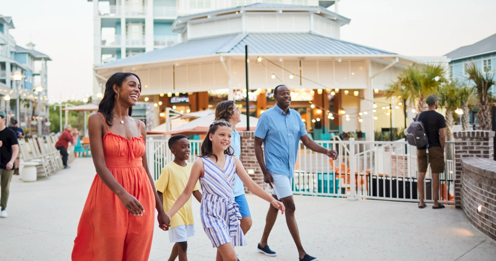 A diverse family walks along a sunny boardwalk near a marina with string lights and a cafe, enjoying a seaside outing together.