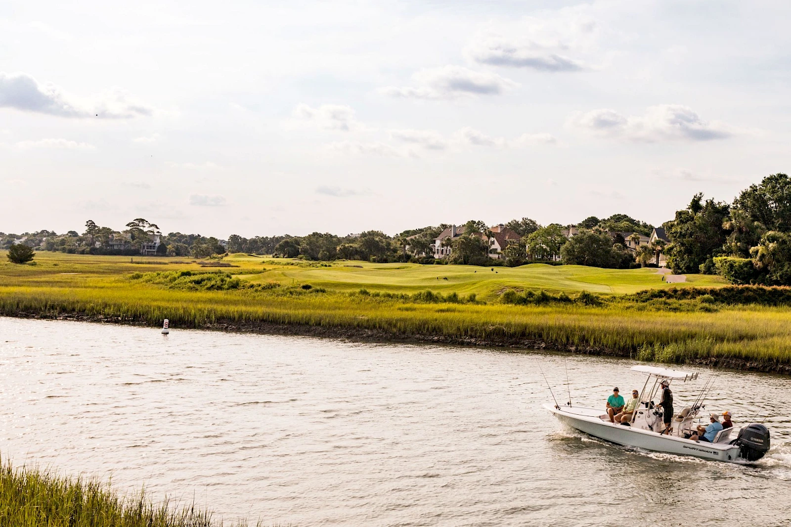 A small boat with people sails along a calm river, a grassy marshland stretches beyond, and distant houses dot the horizon as clouds drift above.