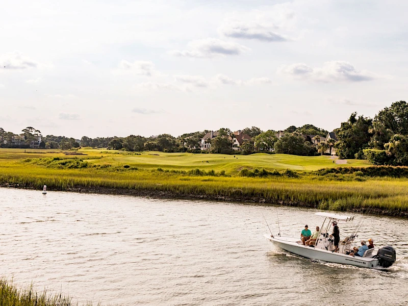 A small boat with people sails along a calm river, a grassy marshland stretches beyond, and distant houses dot the horizon as clouds drift above.