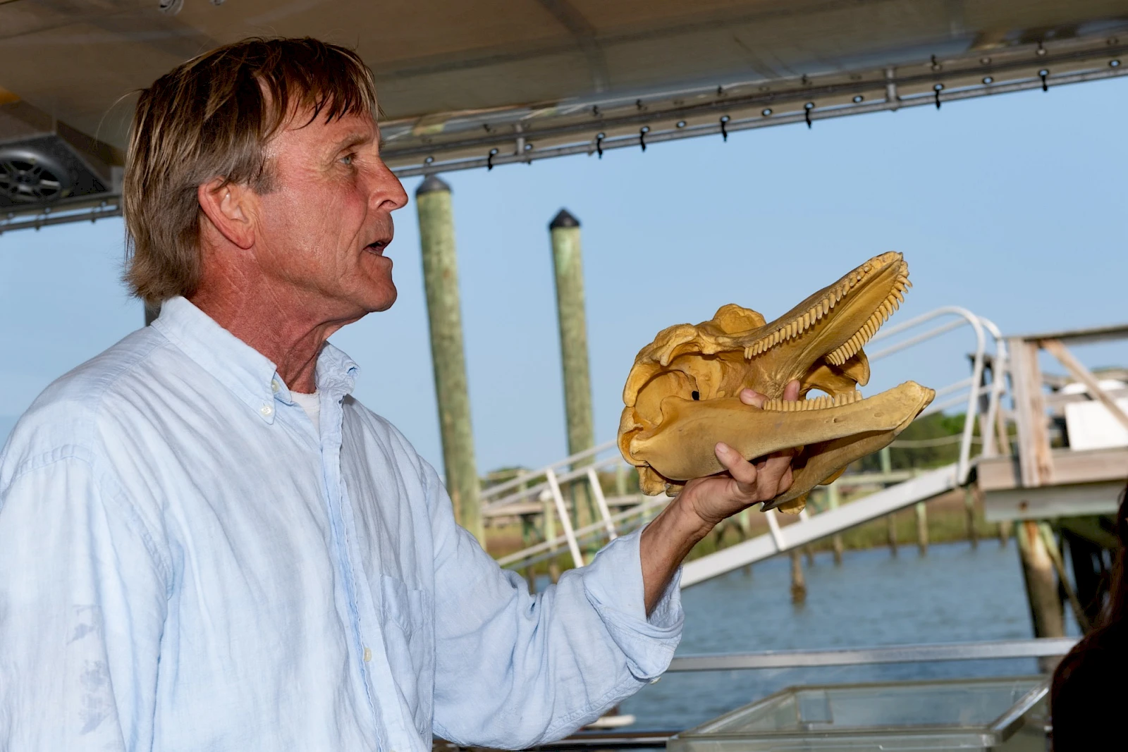 A man in a light blue shirt holds a carved wooden alligator head on a dock, a waterfront setting in the background, mid-speech.