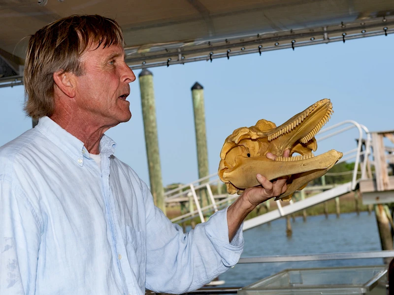 A man in a light blue shirt holds a carved wooden alligator head on a dock, a waterfront setting in the background, mid-speech.