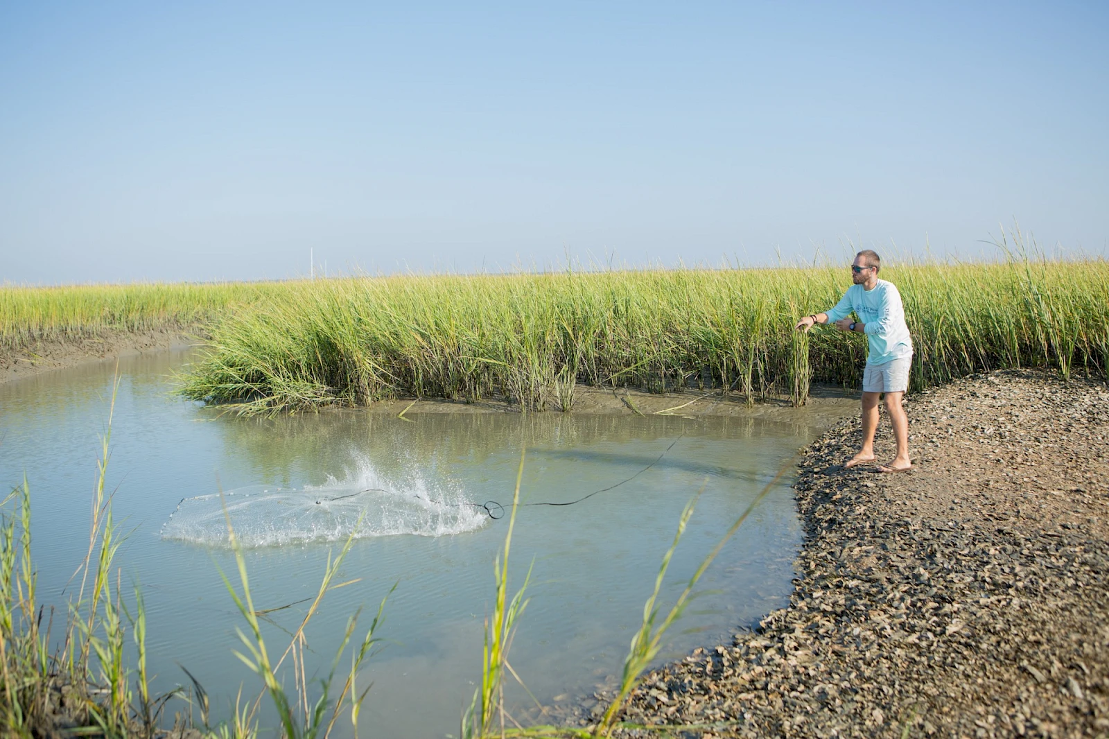 A girl in a blue dress stands on a rocky shore by a calm marsh, fishing or casting into a small pond with tall grasses in the background.