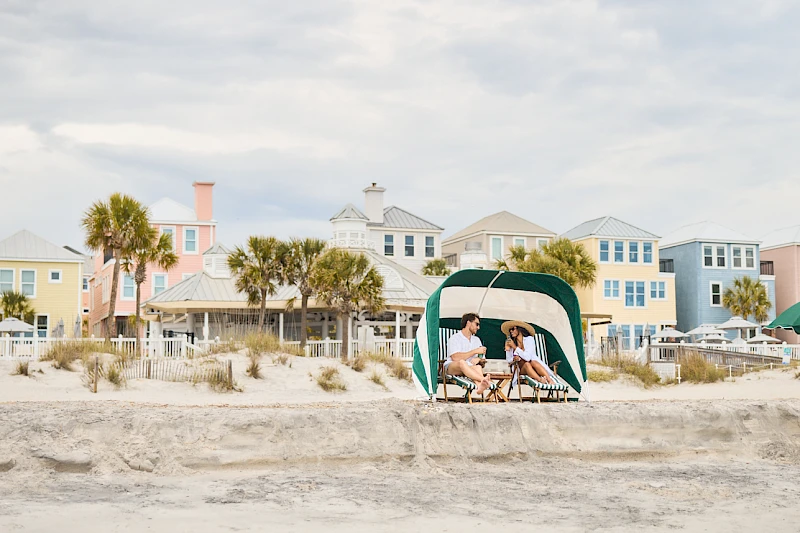 Two people sit under a green beach tent on a sandy shore with colorful houses in the background, enjoying a sunny day at the beach.