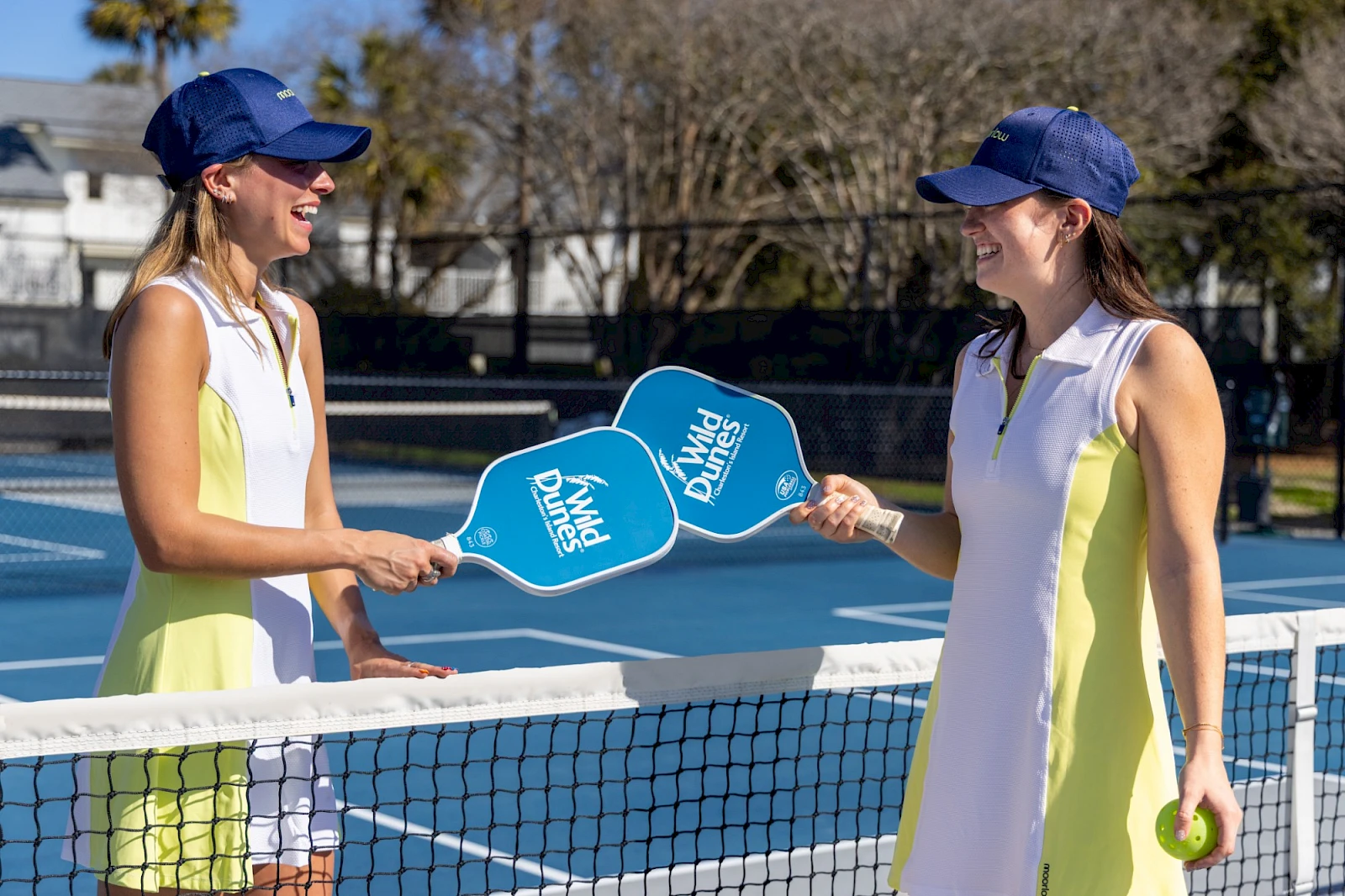 Two women in matching yellow-white dresses play paddle tennis at a net, smiling and shaking paddles by a sunny outdoor court.