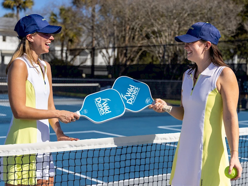 Two women in matching yellow-white dresses play paddle tennis at a net, smiling and shaking paddles by a sunny outdoor court.