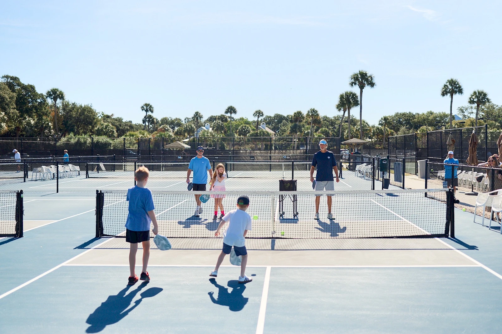 People playing pickleball on outdoor courts, hitting paddles, nets up, sunny day, palm trees in background, several kids and adults active.