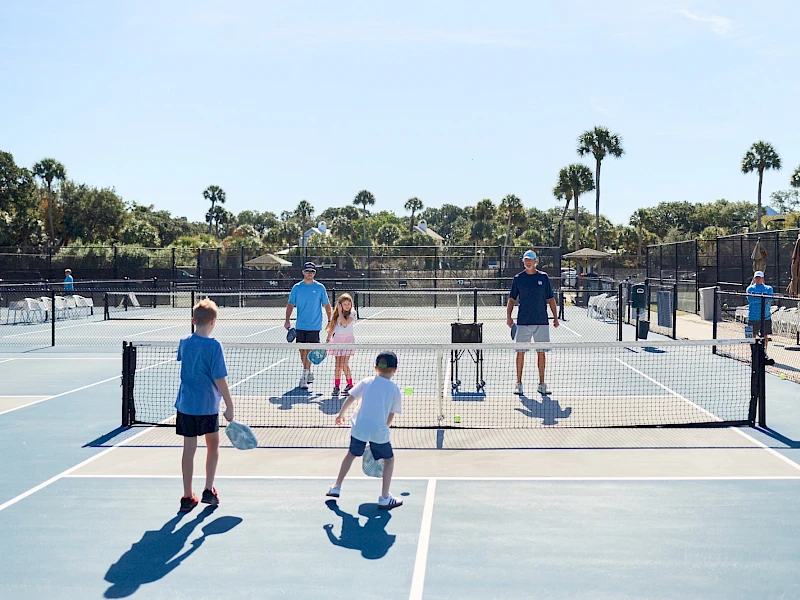 People playing pickleball on outdoor courts, hitting paddles, nets up, sunny day, palm trees in background, several kids and adults active.