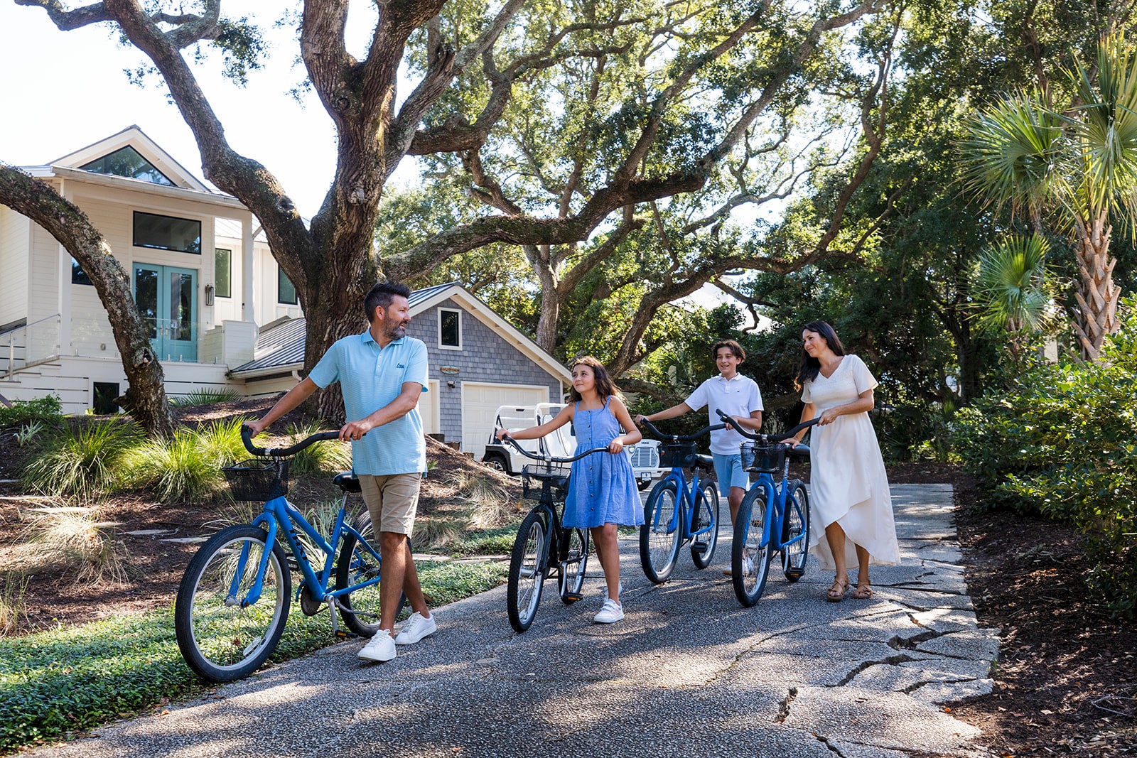 Four people with bikes chat on a sunny residential street, pointing and smiling near a pair of small houses and leafy trees.