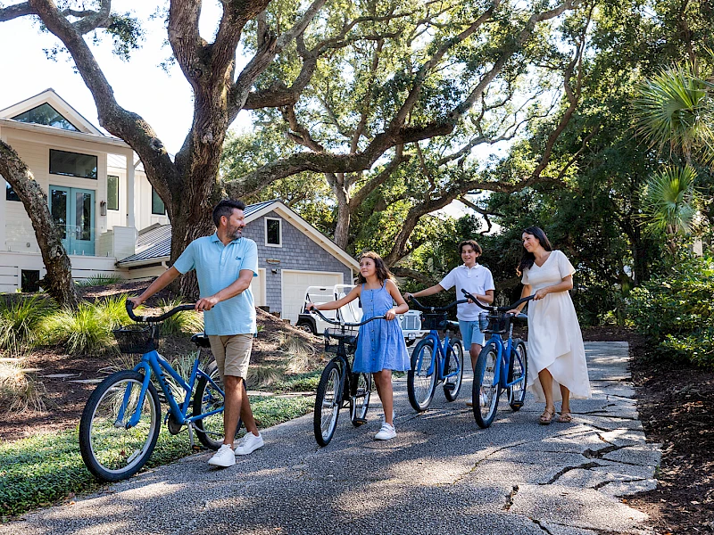 Four people with bikes chat on a sunny residential street, pointing and smiling near a pair of small houses and leafy trees.