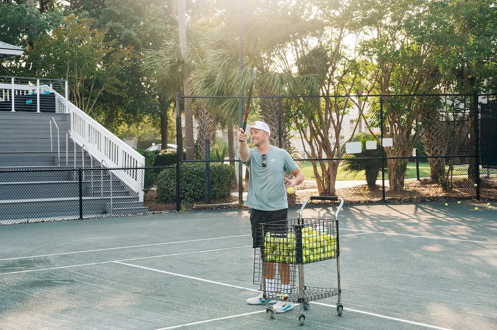 A man with a cap pushes a shopping cart full of tennis balls on an outdoor tennis court, raising one hand as if signaling hello.