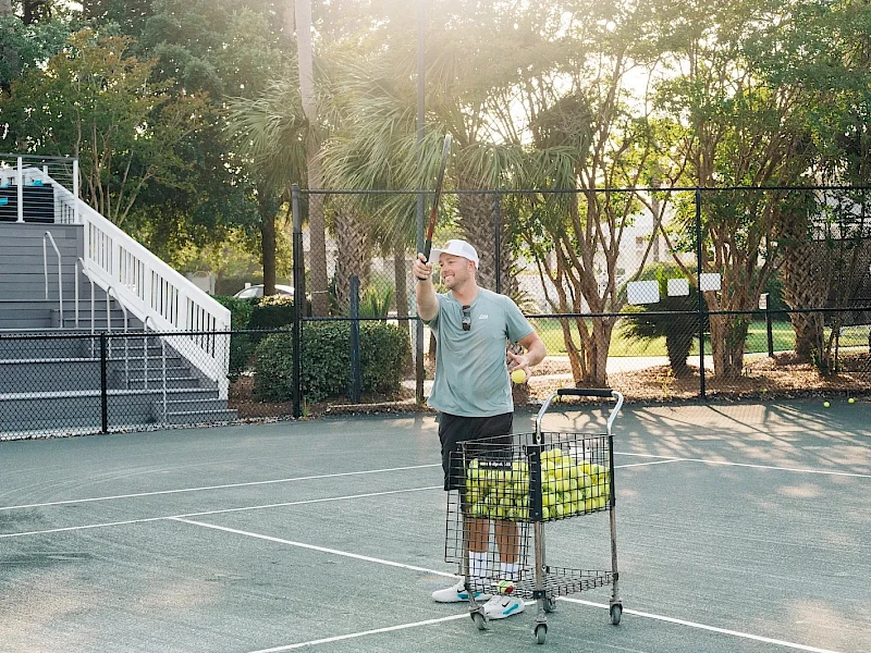 A man with a cap pushes a shopping cart full of tennis balls on an outdoor tennis court, raising one hand as if signaling hello.