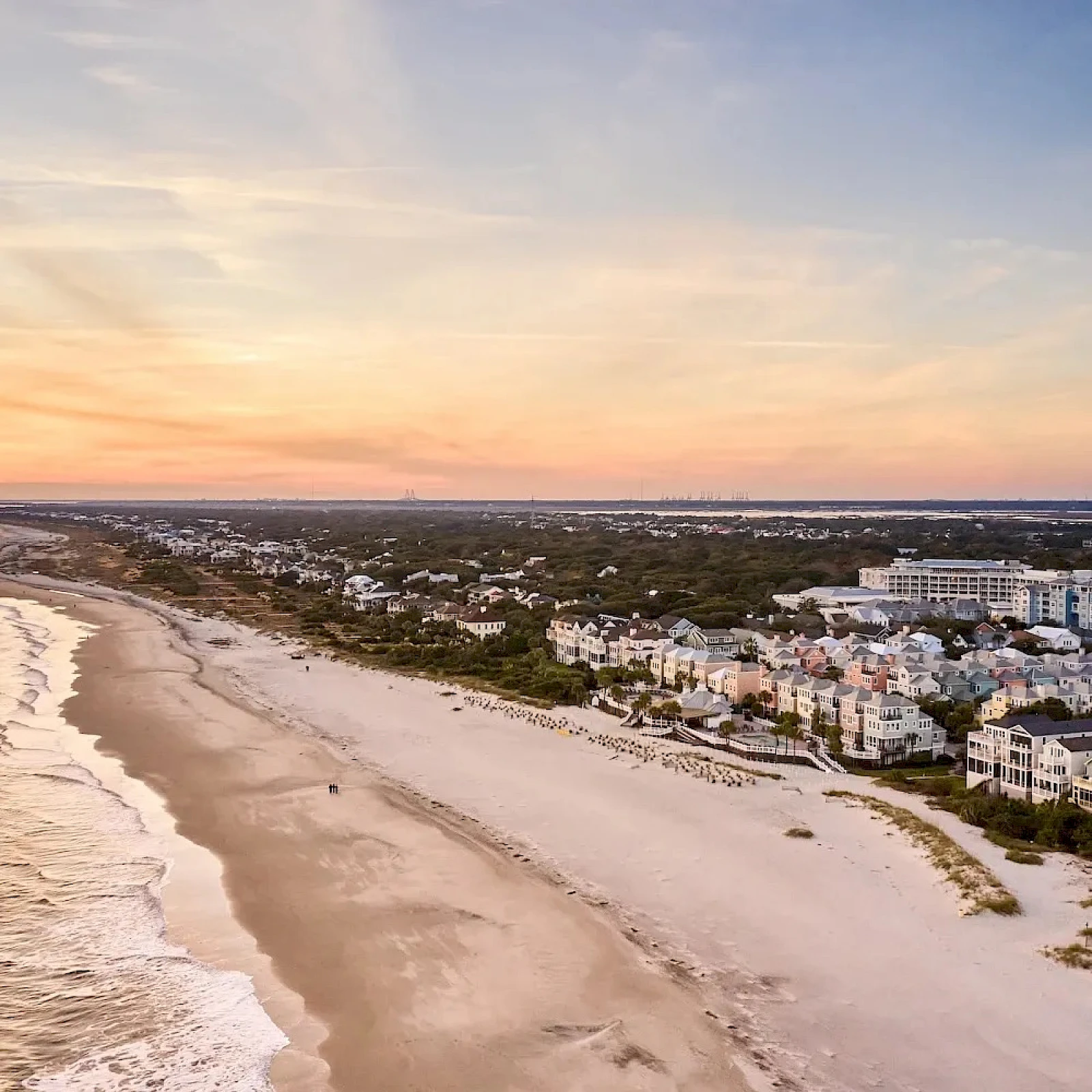 Aerial view of a coastal town at sunset: sandy beach, gentle waves, white buildings lining the shore, and a calm, colorful sky.