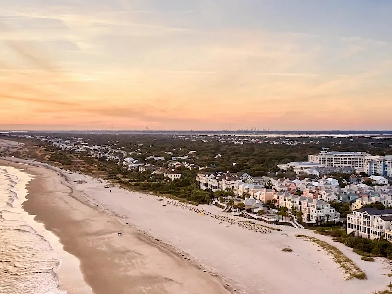 Aerial view of a coastal town at sunset: sandy beach, gentle waves, white buildings lining the shore, and a calm, colorful sky.