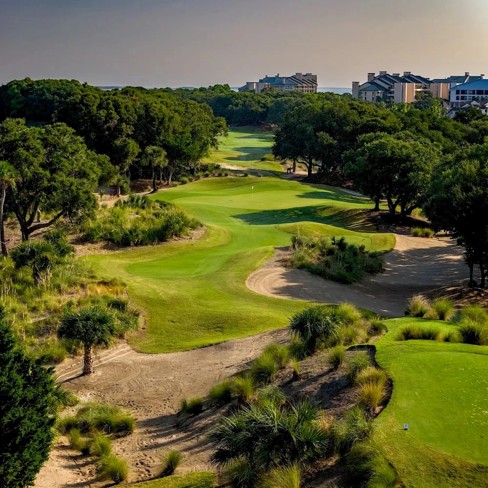 A sunlit golf course with lush greens, winding fairways, sand bunkers, and trees lining the holes, with buildings in the distance.