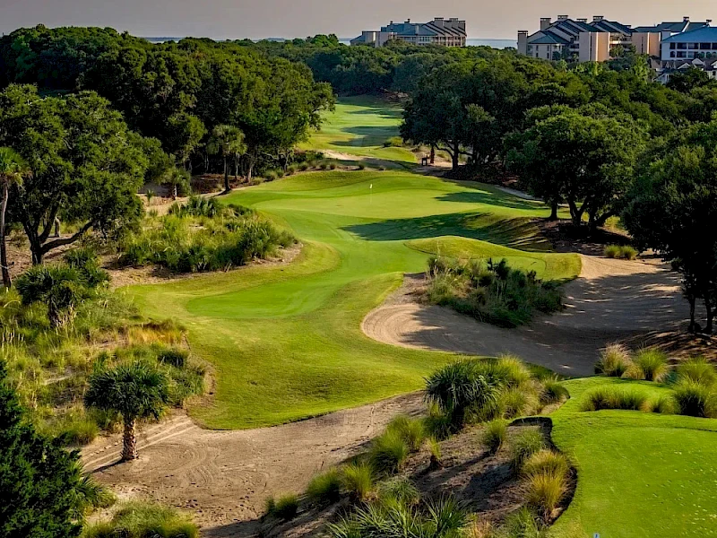 A sunlit golf course with lush greens, winding fairways, sand bunkers, and trees lining the holes, with buildings in the distance.