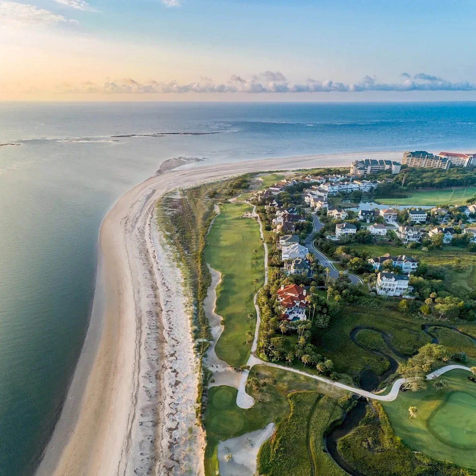 Coastal aerial view of a sandy beach curving along the shoreline, with a green residential area, golf course, and winding pathways by the coast.