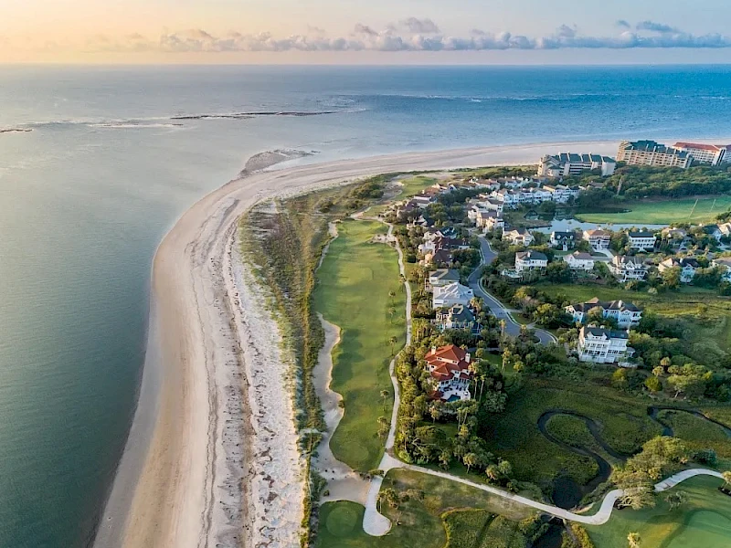 Coastal aerial view of a sandy beach curving along the shoreline, with a green residential area, golf course, and winding pathways by the coast.