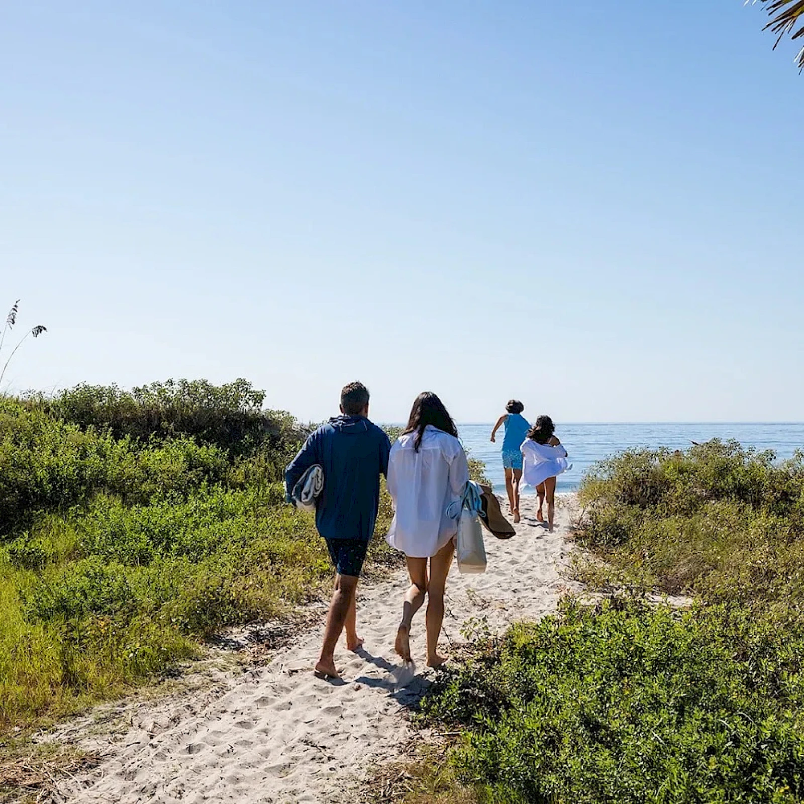 Two people walk hand-in-hand along a sandy coastal path, followed by two others toward the water, with green shrubs and a clear blue sky.