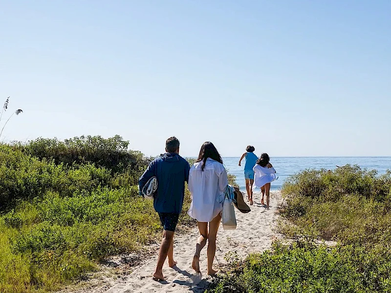 Two people walk hand-in-hand along a sandy coastal path, followed by two others toward the water, with green shrubs and a clear blue sky.