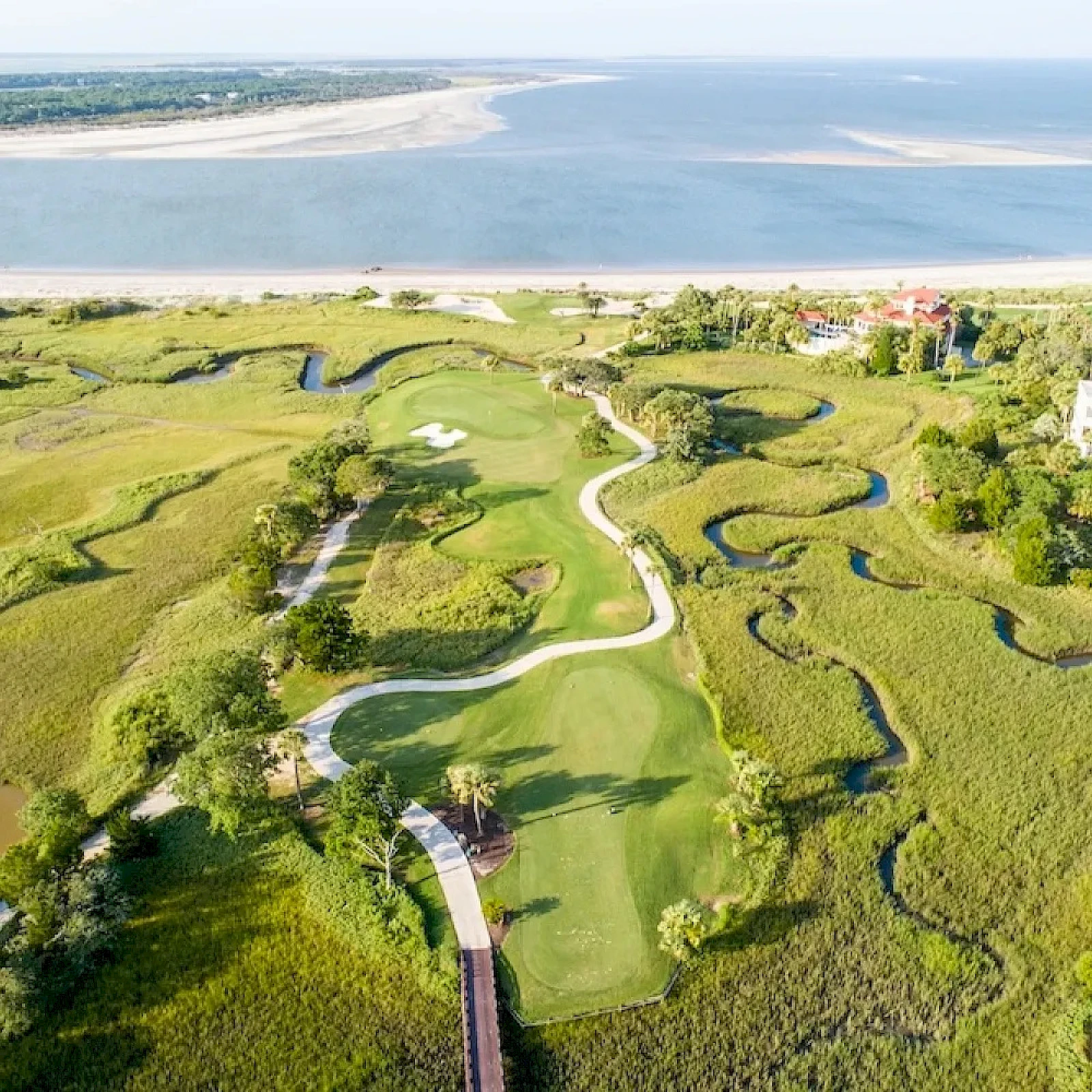 Aerial view of a coastal golf course with winding greens, fairways, and water hazards, bordered by dunes and a beach beyond.
