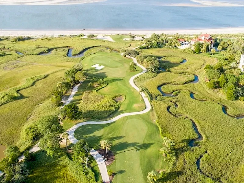 Aerial view of a coastal golf course with winding greens, fairways, and water hazards, bordered by dunes and a beach beyond.