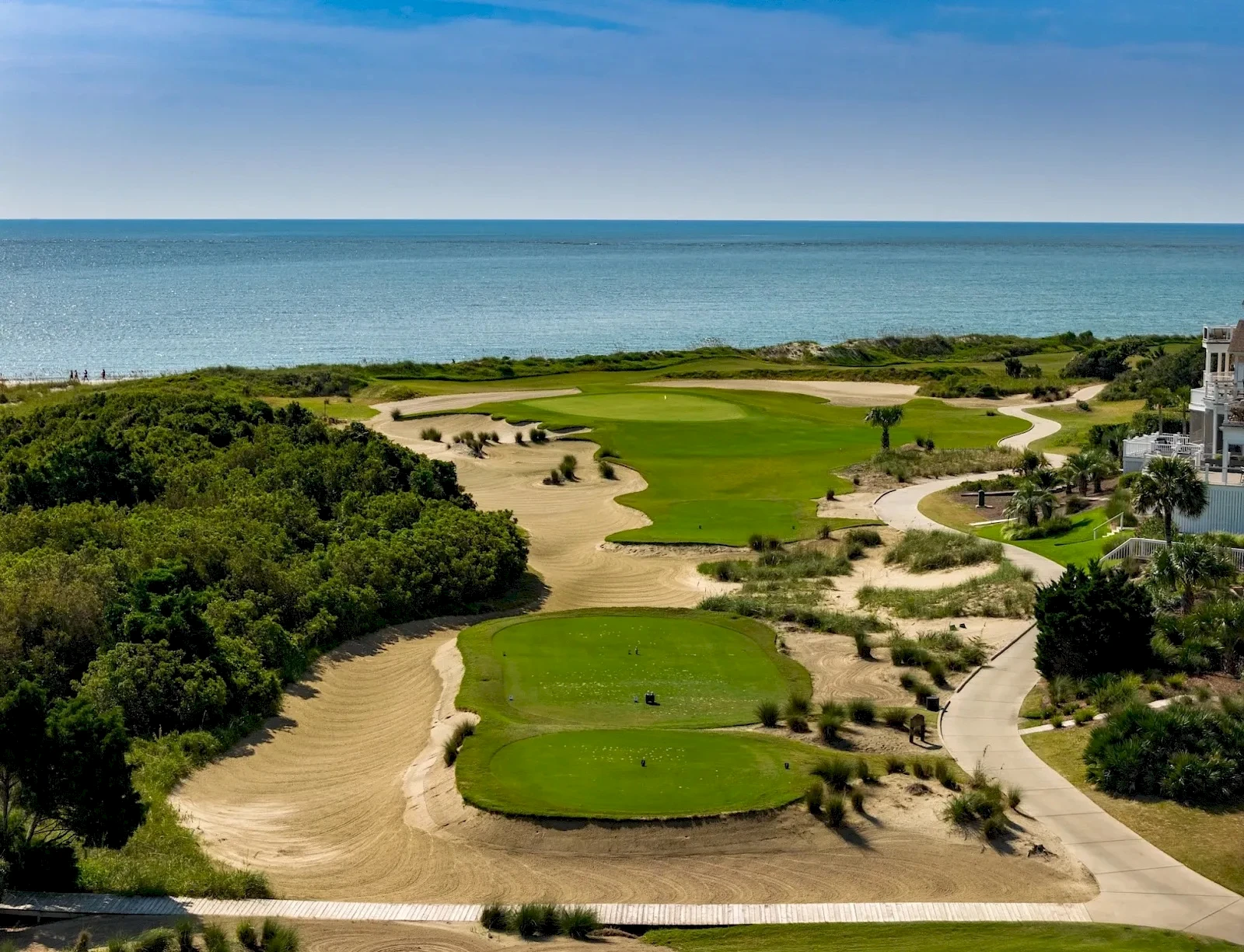 A coastal golf course with green fairways, sandy bunkers, and paths weaving toward the sea under a blue sky.