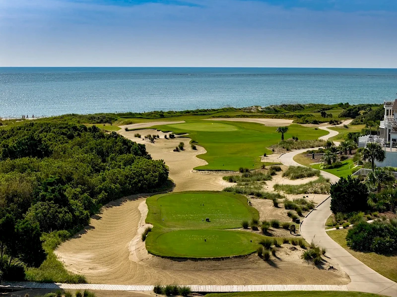 A coastal golf course with green fairways, sandy bunkers, and paths weaving toward the sea under a blue sky.