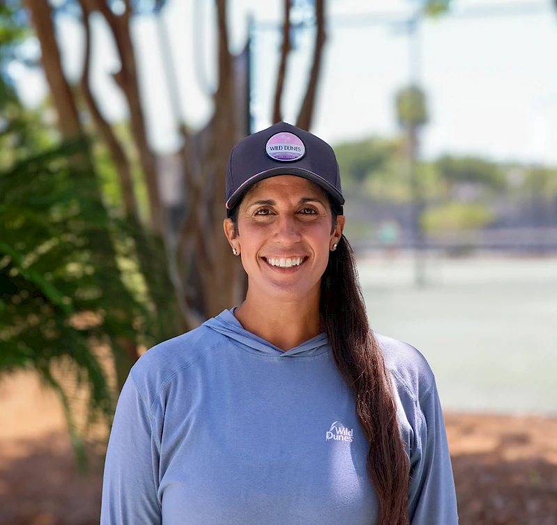 A smiling woman in a blue hoodie and cap stands outdoors with trees in the background, looking at the camera.