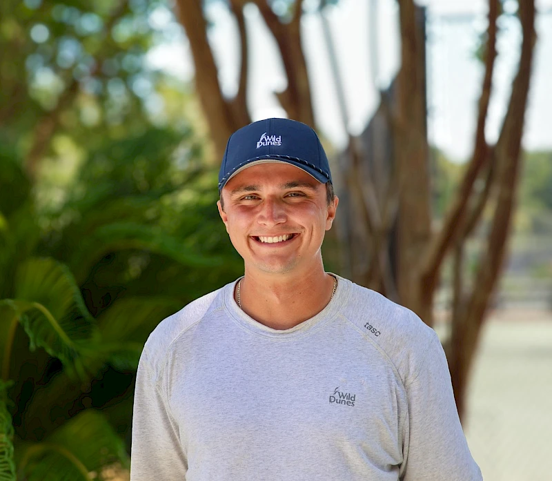 A smiling person in a light gray t-shirt and blue cap stands outdoors near trees, sunny day and tropical plants in the background.