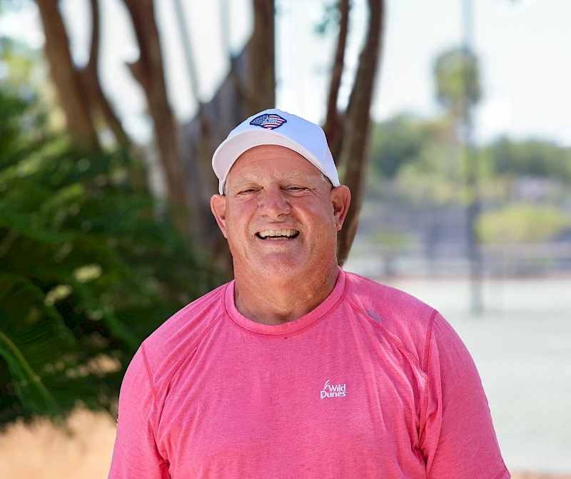A smiling man in a pink athletic shirt and white cap standing outdoors, with trees and a fence in the background.