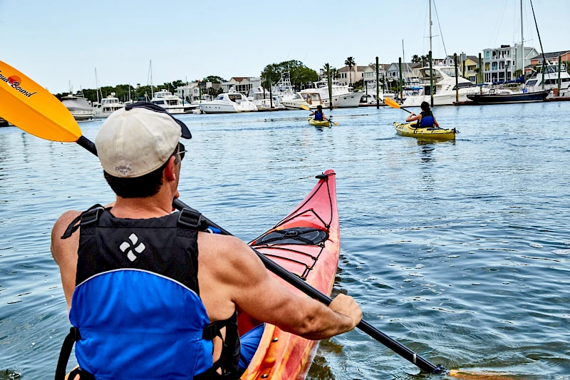A man in a blue life vest kayaks on calm water, paddling toward a marina with sailboats and houses in the background, sunny weather.
