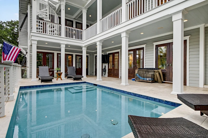 A modern two-story house with a rectangular backyard pool, white columns, lounge chairs, a porch, and an American flag—vacation vibes abound.