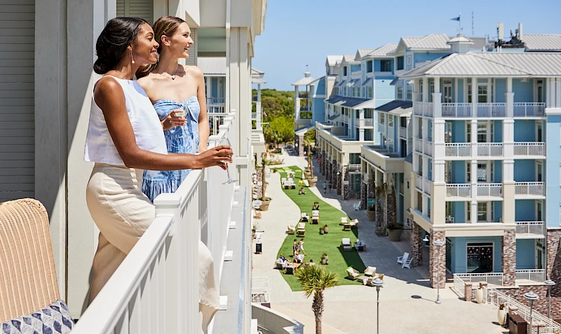 Two women smile and chat on a balcony overlooking a sunny resort with pools and row of modern buildings below.