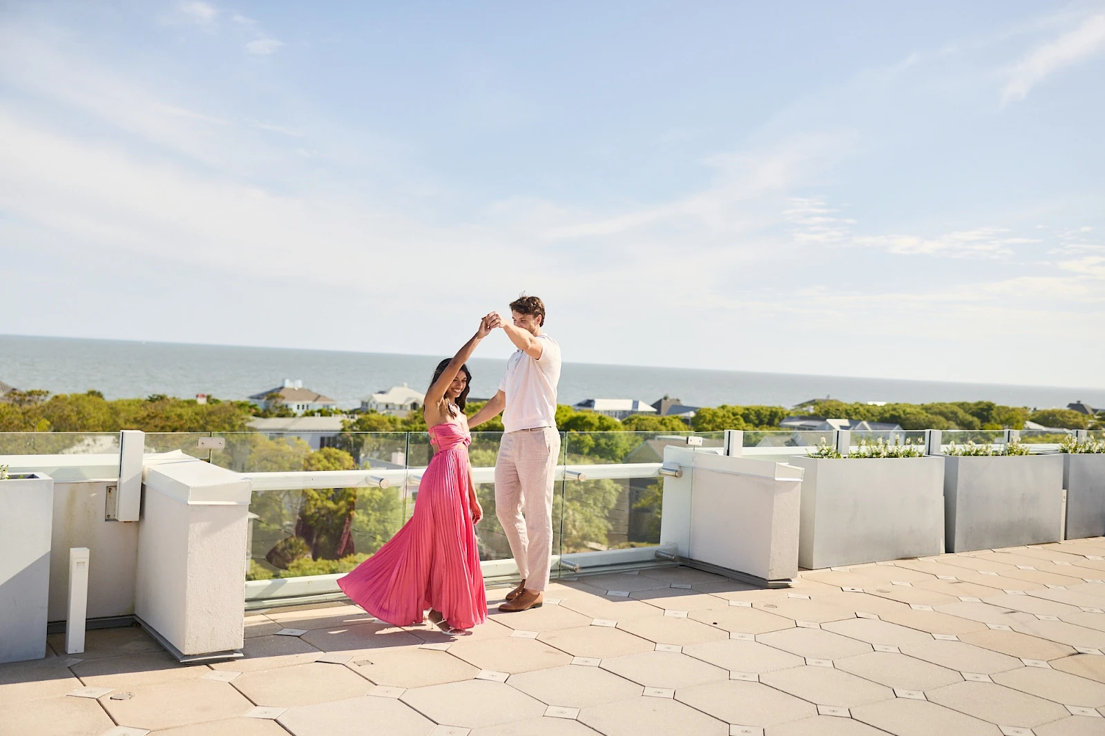 Two people dance on a sunny terrace overlooking the sea; a woman in a pink dress frames a man in light clothing as they twirl together.