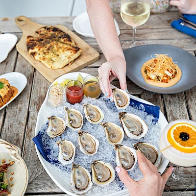 Assorted appetizers and dishes on a rustic table: oysters on a platter, spoons of sauces, a pumpkin soup, a small pasta plate, a flatbread, and a glass of white wine.