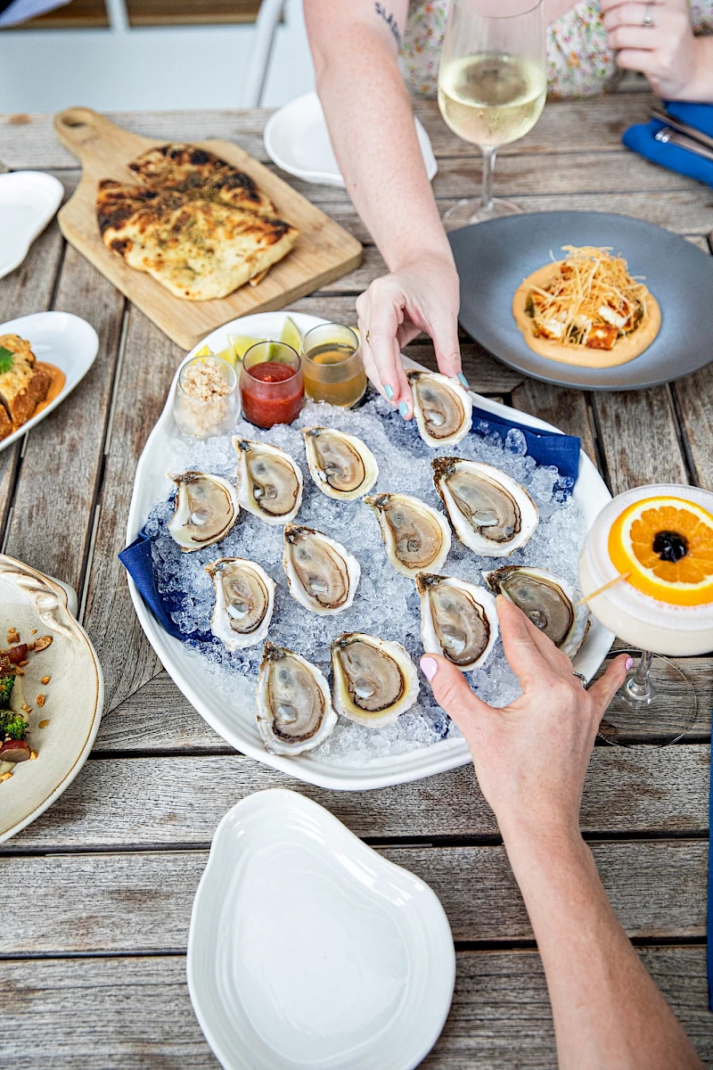 Assorted appetizers and dishes on a rustic table: oysters on a platter, spoons of sauces, a pumpkin soup, a small pasta plate, a flatbread, and a glass of white wine.
