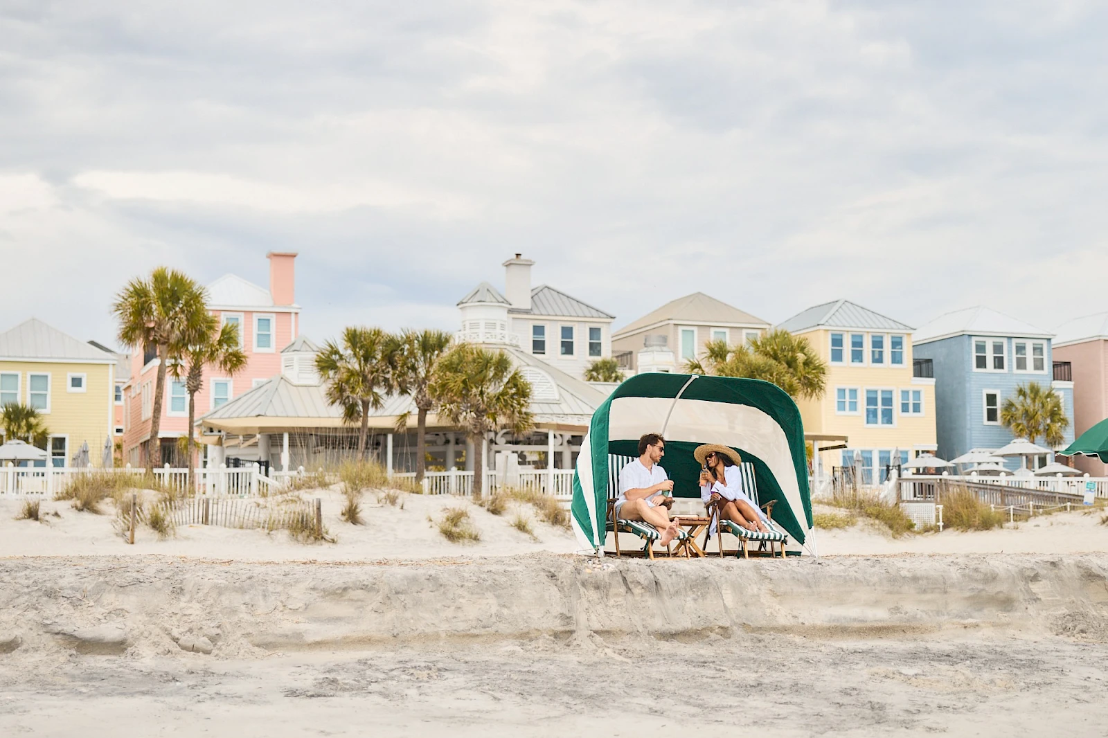 Two people sit under a green canopy on a sandy beach, chatting, with colorful pastel houses lining the background and a calm, sunny sky.