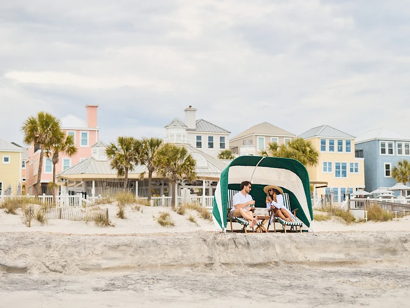 Two people sit under a green canopy on a sandy beach, chatting, with colorful pastel houses lining the background and a calm, sunny sky.