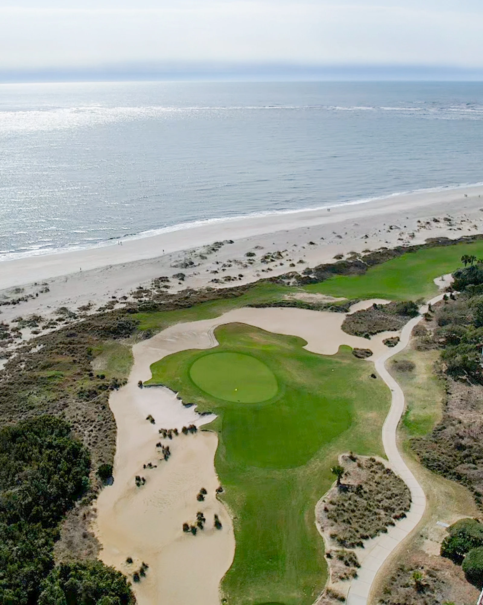 An aerial view of a beach golf course with greens, fairways, sand traps, and surrounding dunes along the shoreline, under a hazy sky.